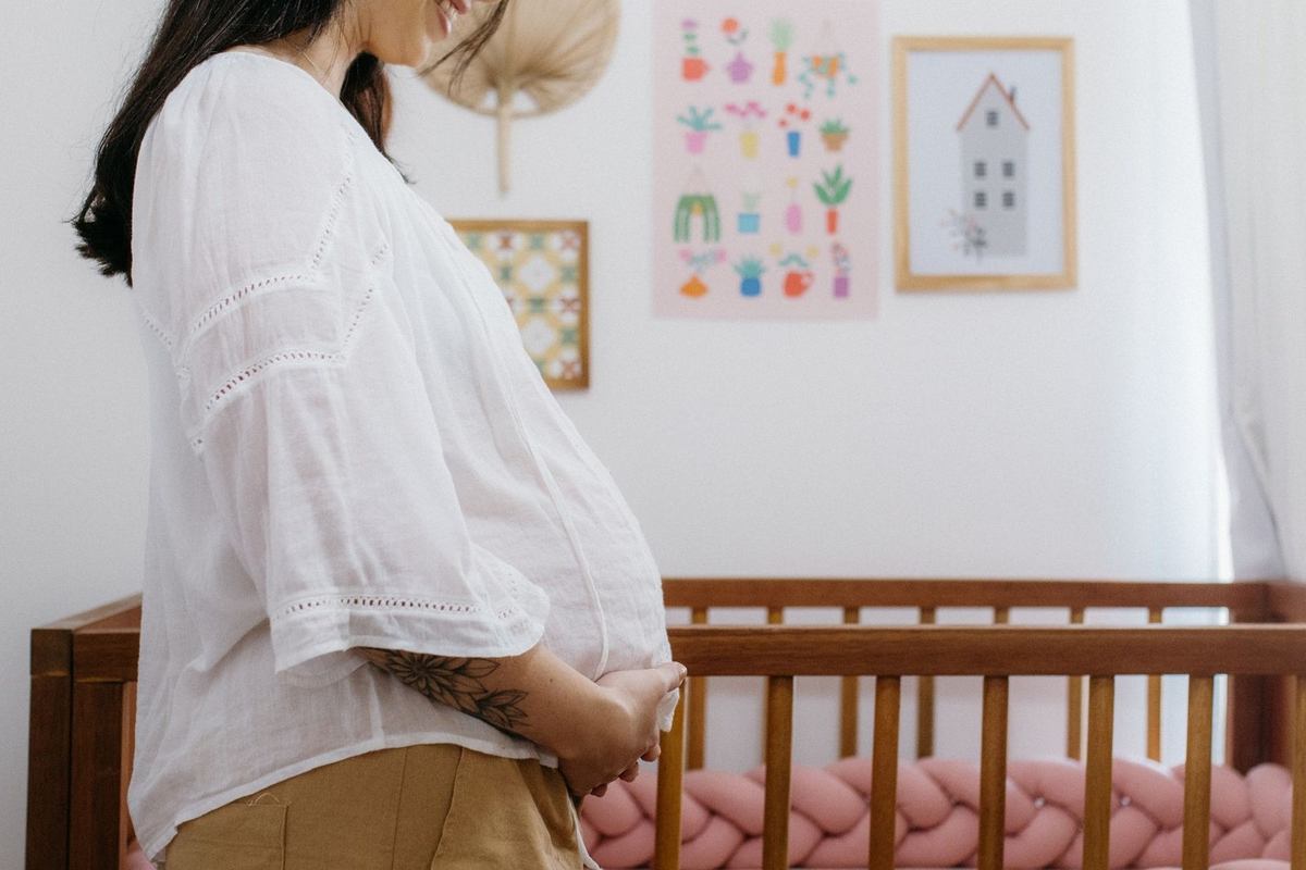 Femme enceinte dans une chambre, main sur ventre, lit d'enfant avec coussin tressé, oeuvres murales Femme enceinte dans une chambre, main sur ventre, lit d'enfant avec coussin tressé, oeuvres murales